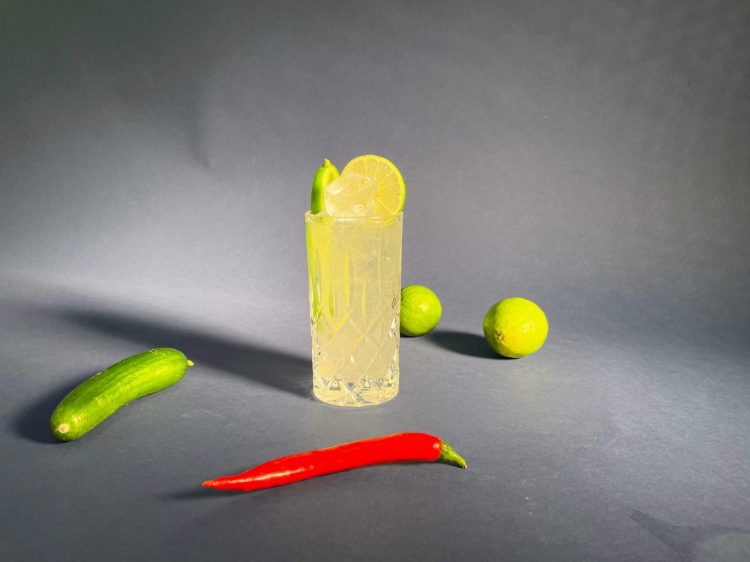 Das Bild zeigt einen Longdrink mit Gurke, Chilis und Limetten im Hintergrund. Es veranschaulicht das Moscow Mule Cocktail Rezept.